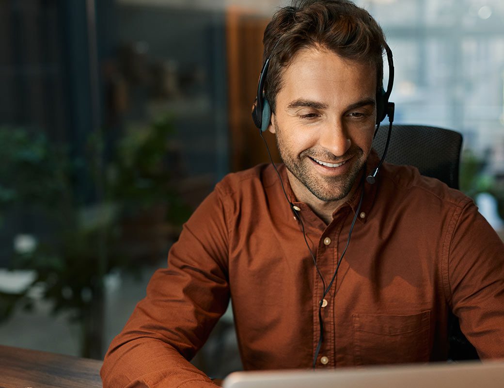 Young man sits, smiling, at his laptop wearing headphones and an orange dress shirt.